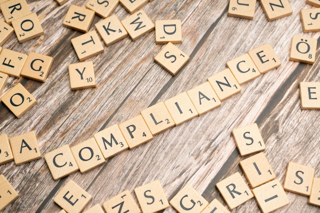 Wooden Scrabble tiles spelling out 'COMPLIANCE' on a rustic wood surface, surrounded by scattered letter tiles